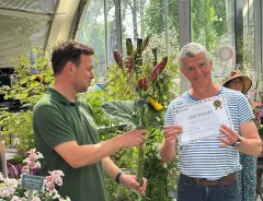 Two men in a greenhouse hold a certificate beside colorful flower arrangements.