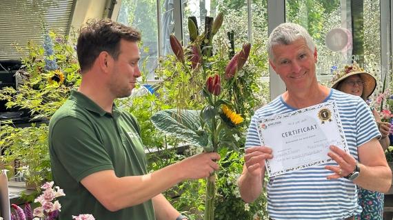 Two men in a greenhouse hold a certificate beside colorful flower arrangements.
